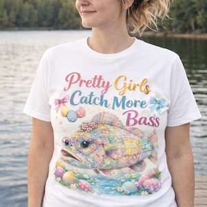Może przedstawiać: Biała koszulka z napisem "Pretty Girls Catch More Bass" w kolorowych literach. Koszulka przedstawia fantazyjną ilustrację ryby w okularach w kształcie serca i kwiatowej koronie, otoczonej pisankami.