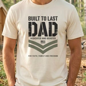 May include: A beige t-shirt with the text "BUILT TO LAST DAD" in black, with "DEDICATED AND DEVOTED" below. A small American flag and olive green chevron design are also present. The phrase "FOR FAITH, FAMILY AND FREEDOM" is at the bottom.