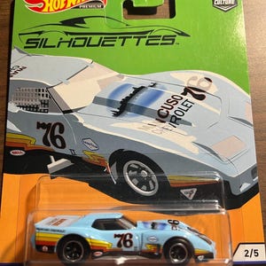 Könnte beinhalten: Ein Spielzeugauto '76 Greenwood Corvette in einer transparenten Kunststoffverpackung, Teil der Hot Wheels Car Culture-Serie. Das Auto ist hellblau mit der Nummer 76 auf der Seite und oben. Die Verpackung ist grün mit dem Hot Wheels-Logo.