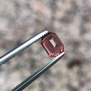 May include: A rectangular, rose-coloured gemstone is held by silver tweezers. The gem has a faceted surface and a polished appearance. The background is a blurred, neutral tone. The gem's edges are sharp and well-defined.