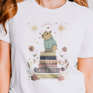 May include: White t-shirt featuring a whimsical illustration of a capybara wearing a sweater and glasses, sitting atop a stack of books. The design includes floral and celestial elements in soft colors.