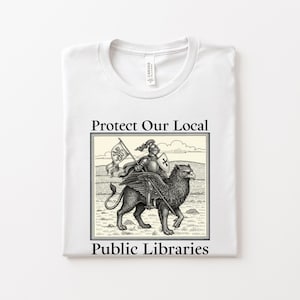 May include: White t-shirt with a graphic that reads "Protect Our Local Public Libraries." The graphic features a knight riding a griffin, holding a flag with a heraldic design. The design is in black and white.