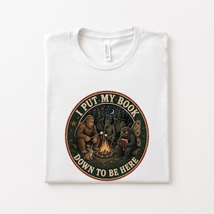 Può includere: Una t-shirt bianca con una grafica circolare che riporta il testo I PUT MY BOOK DOWN TO BE HERE. L'illustrazione raffigura una scena attorno a un falò con un sasquatch, una creatura alata e un essere simile a una falena in un bosco.