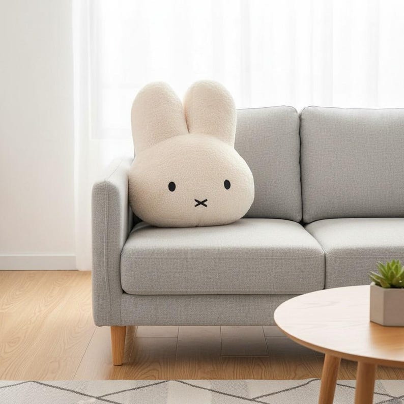 May include: A cream-colored bunny-shaped pillow with black eyes and a stitched mouth rests on a gray sofa. The plush pillow has large, upright ears and a soft, textured surface. The pillow adds a touch of whimsy to the living room setting.