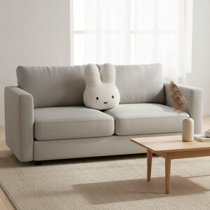 May include: A light gray two-seater sofa with a white bunny-shaped pillow. The pillow has black eyes and a black cross for a mouth. A small wooden coffee table and a vase with dried plants are in front of the sofa.