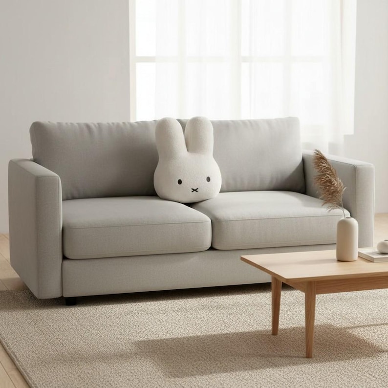 May include: A light gray two-seater sofa with a white bunny-shaped pillow. The pillow has black eyes and a black cross for a mouth. A small wooden coffee table and a vase with dried plants are in front of the sofa.
