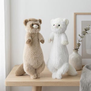 May include: Two plush ferret stuffed animals. One is light brown with darker brown accents, the other is white. Both have small black eyes and stand upright on a wooden table. The background is a neutral color.