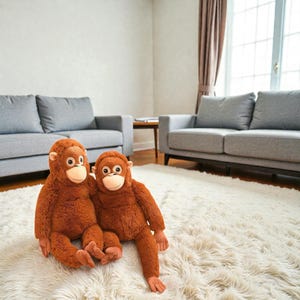 May include: Two plush orangutan stuffed animals, with brown fur and tan faces, sit on a white shag rug. The stuffed animals have large eyes and are positioned in a living room setting with gray sofas and a window.