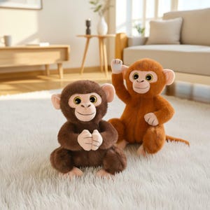 Può includere: Due scimmie di peluche. Una è marrone scuro, con le mani giunte. L'altra è marrone chiaro, con un braccio alzato. Entrambe hanno grandi occhi e sono sedute su un tappeto bianco e soffice. Giocattoli per bambini.