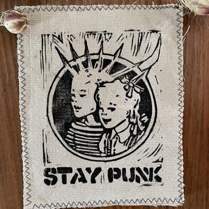 Könnte beinhalten: Ein kleines, rechteckiges Stoffpatch mit einem schwarz-weißen Linolschnitt. Der Druck zeigt zwei stilisierte Figuren in einem Kreis, darunter der Text "STAY PUNK". Das Patch hat einen rohen, genähten Rand und ist vor einem Holzgrund platziert.