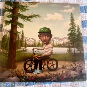 May include: Album cover art featuring a person on an orange bicycle with black wheels. The person wears a green cap with "WOLF" on it and a striped shirt. The background shows a lake, mountains, and trees. A bee flies near a tree.