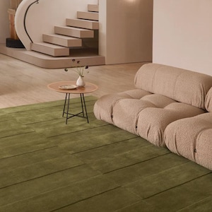 Può includere: Un tappeto verde oliva con un design lineare strutturato copre il pavimento. Un tavolino basso e rotondo con base in metallo scuro e piano marrone chiaro è al centro. Un divano beige e una scala curva sono sullo sfondo.
