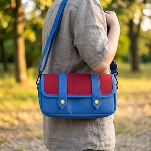 Puede incluir: Bolso mensajero azul y rojo con una correa para el hombro azul. El bolso tiene forma rectangular con solapa roja y cuerpo azul. Botones amarillos aseguran la solapa. El bolso se lleva al hombro.