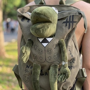May include: An olive green backpack with a plush frog attached. The backpack has a zip closure and adjustable straps. The frog has a white and black vest, and the backpack has doodles and the text "No A".