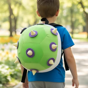 Puede incluir: Una mochila verde y blanca con un diseño puntiagudo, con detalles morados y crema. La mochila la lleva una persona con una camisa azul, caminando al aire libre.