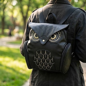 Puede incluir: Mochila negra con forma de búho con detalles bordados dorados. La mochila presenta una solapa que forma la cara del búho, con detalles dorados alrededor de los ojos y un patrón bordado dorado en la parte delantera. Tiene dos bolsillos laterales y correas ajustables.
