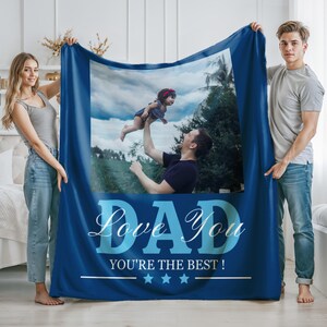 May include: A dark blue fleece blanket featuring a large central photograph of a person lifting a toddler into the air against a cloudy sky. The text DAD LOVE YOU YOU'RE THE BEST! is printed in light blue and white below the photo with three stars.
