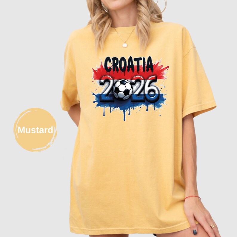 May include: Mustard-colored t-shirt featuring the word "CROATIA" above the numbers "2026" with a soccer ball in the center. The design incorporates red, white, and blue accents, and the shirt has a relaxed fit.