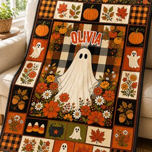 May include: A rectangular quilt featuring a central ghost graphic with the name OLIVIA printed above it. The design includes a black and white buffalo check pattern, orange pumpkins, autumn leaves, flowers, acorns, candy corn, and a black cauldron.