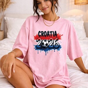 May include: A light pink t-shirt with the word "CROATIA" in black, above the numbers "2026" and a soccer ball graphic. The design has red, white, and blue accents. The shirt is worn by a person.