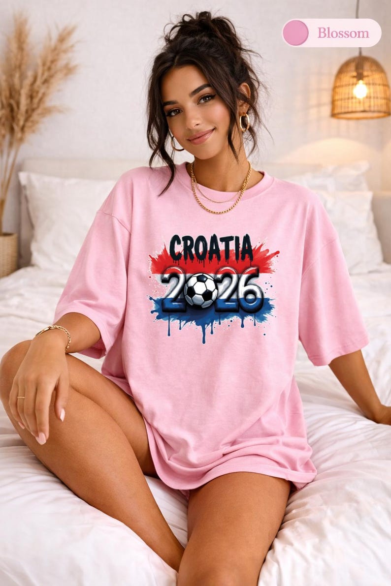 May include: A light pink t-shirt with the word "CROATIA" in black, above the numbers "2026" and a soccer ball graphic. The design has red, white, and blue accents. The shirt is worn by a person.