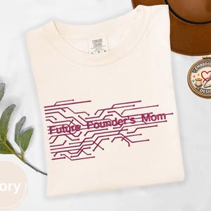 May include: Ivory-colored t-shirt with the text "Future Founder's Mom" in a burgundy circuit board design. The shirt is a Comfort Colors brand, with a tag indicating size L-G-G. A brown hat and an embroidered design are in the background.