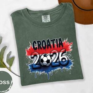 May include: Moss green t-shirt with the word "CROATIA" in black, and the numbers "2026" in white and blue, with a soccer ball in the center. The design has red and blue paint splatter accents. The shirt is folded.