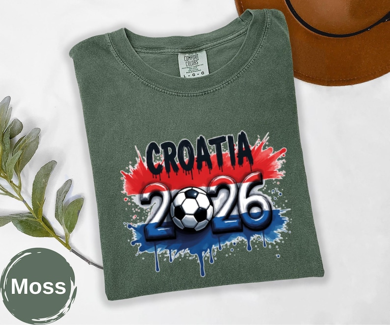 May include: Moss green t-shirt with the word "CROATIA" in black, and the numbers "2026" in white and blue, with a soccer ball in the center. The design has red and blue paint splatter accents. The shirt is folded.
