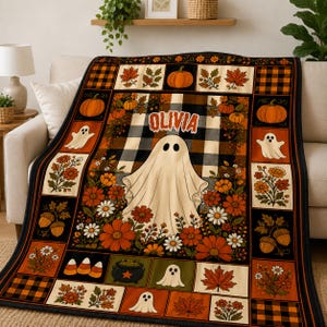 May include: A rectangular throw blanket featuring a central white ghost and the name OLIVIA in orange letters. The design includes a black and white buffalo plaid background, orange pumpkins, autumn leaves, flowers, acorns, and candy corn motifs.
