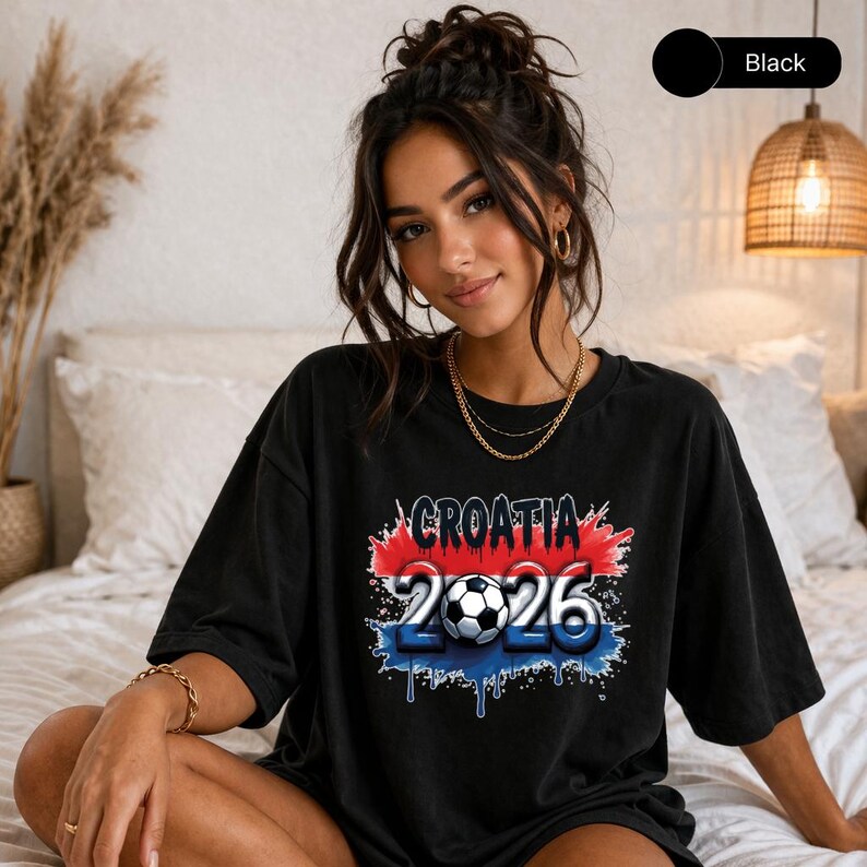 May include: Black t-shirt featuring the text "CROATIA 2026" with a soccer ball graphic. The text is in white, red, and blue, with a distressed paint effect. The shirt is a casual, oversized style.