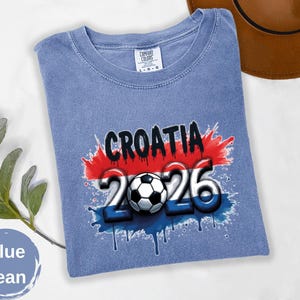 May include: Blue jean t-shirt with a graphic design featuring the word "CROATIA" in black, the number "2026" in white and blue, and a soccer ball. The design incorporates the colors of the Croatian flag.