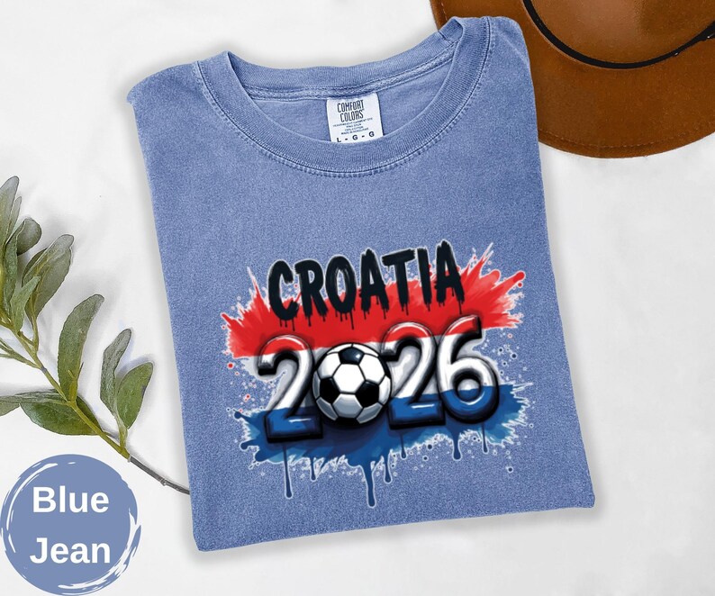 May include: Blue jean t-shirt with a graphic design featuring the word "CROATIA" in black, the number "2026" in white and blue, and a soccer ball. The design incorporates the colors of the Croatian flag.