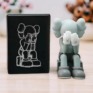 May include: A light green figurine of a cartoon character with hands covering its face, wearing black shorts and grey boots. Beside it is a black box with a white illustration of the same character.