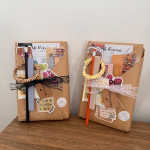 Può includere: Due pacchi di carta marrone legati con nastri e decorati con adesivi, matite ed elementi decorativi. Un pacco ha un nastro nero e l'altro un elastico giallo. I pacchi sono etichettati "Romance" e "Book Review".