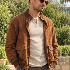 Puede incluir: Una chaqueta bomber de ante marrón con cuello y puños acanalados, con cierre de botones, dos bolsillos delanteros con solapa y un polo beige claro debajo, combinado con pantalones gris oscuro y gafas de sol de carey.