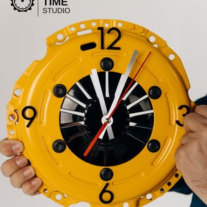 May include: A circular wall clock crafted from a yellow automotive clutch pressure plate. The clock features black numerals at the 12, 3, 6, and 9 positions, a black center with white and silver hands, and a thin red second hand.