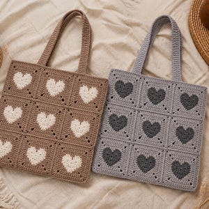 May include: Two crochet tote bags. One is brown with white hearts, the other is gray with dark gray hearts. Each bag has a square design with nine heart-shaped patterns. The bags have matching crochet handles.