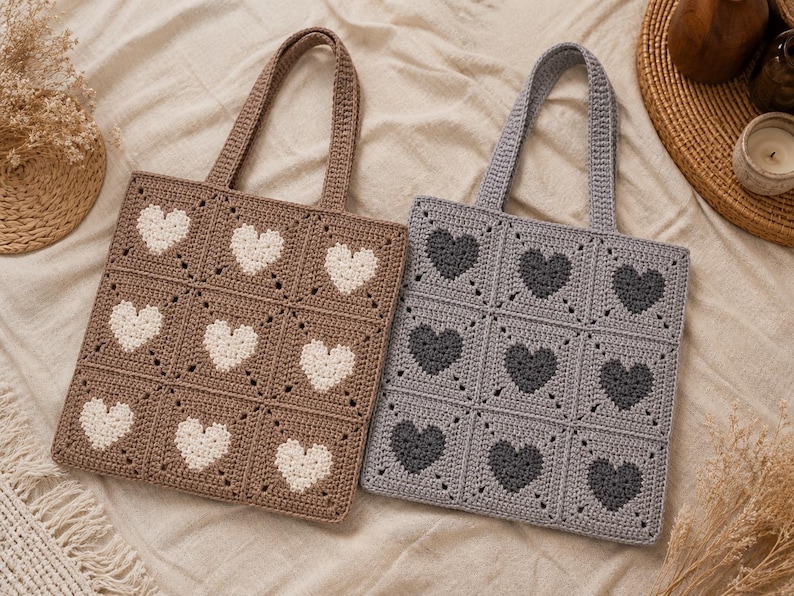 May include: Two crochet tote bags. One is brown with white hearts, the other is gray with dark gray hearts. Each bag has a square design with nine heart-shaped patterns. The bags have matching crochet handles.