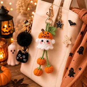 May include: A cream-colored tote bag adorned with Halloween-themed keychains and pins. The keychains include a black pom-pom, a black cat, and a crochet ghost wearing a pumpkin hat with dangling orange pumpkins. A scarf with ghost and star patterns is draped over the bag.