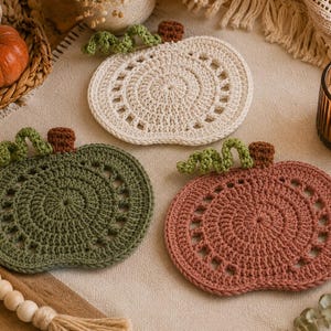 May include: Three crocheted pumpkin-shaped coasters in cream, green, and dusty rose. Each coaster features a spiral design and is topped with a brown stem and green vine. A small orange pumpkin and a lit candle are also visible.