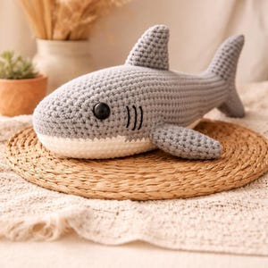 May include: Handmade crochet shark in gray with a white belly, black eye, and stitched gills. The shark is displayed on a woven mat. The text "Handmade Crochet Shark" is written below the shark.