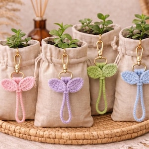 May include: Four small beige drawstring bags, each holding a small plant. Each bag has a gold keychain with a crocheted leaf design in pink, purple, green, and blue. The bags sit on a woven tray.