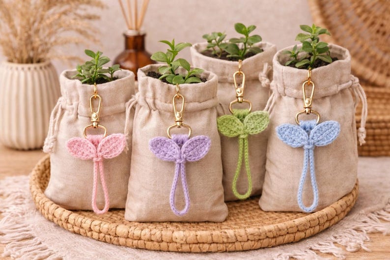 May include: Four small beige drawstring bags, each holding a small plant. Each bag has a gold keychain with a crocheted leaf design in pink, purple, green, and blue. The bags sit on a woven tray.