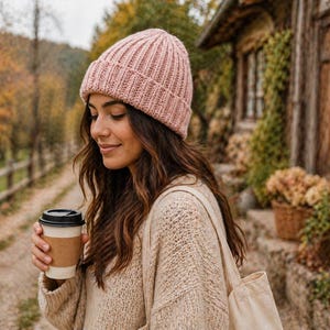 May include: A woman wearing a light pink knit beanie and a beige sweater, holding a coffee cup with a black lid. The background features a rustic building and autumn foliage, creating a cozy, seasonal aesthetic.
