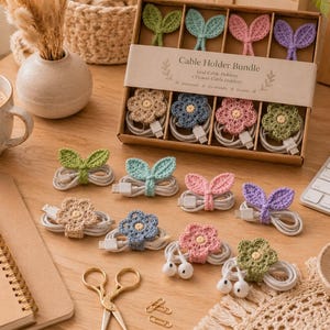 May include: A set of crochet cable holders in a cardboard box labeled Cable Holder Bundle. The set includes various leaf and flower shapes in pastel colors like green, blue, pink, and beige, designed to wrap around white charging cables and cords.