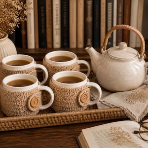 May include: A set of four white ceramic mugs, each encased in a beige crocheted cozy featuring a single wooden button, arranged on a rectangular woven rattan tray alongside a matching speckled ceramic teapot with a curved rattan handle.
