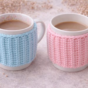 May include: Two white ceramic mugs filled with a light brown beverage, each wrapped in a knitted cozy. One cozy is light blue, the other is pink. The mugs have white handles and floral patterns inside.