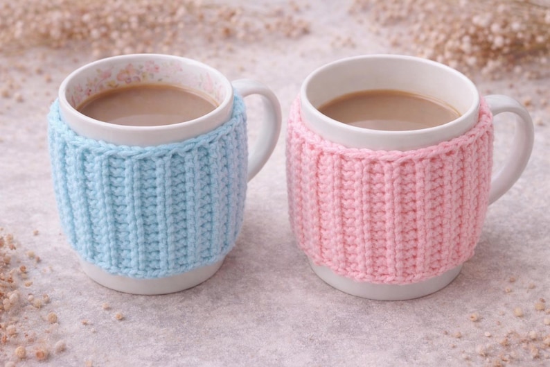 May include: Two white ceramic mugs filled with a light brown beverage, each wrapped in a knitted cozy. One cozy is light blue, the other is pink. The mugs have white handles and floral patterns inside.
