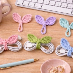 May include: A collection of colorful, handmade crochet cable organizers shaped like leaves. The organizers are in pink, purple, green, and blue, each wrapped around a white charging cable. A keyboard, coffee cup, and pen are also visible.