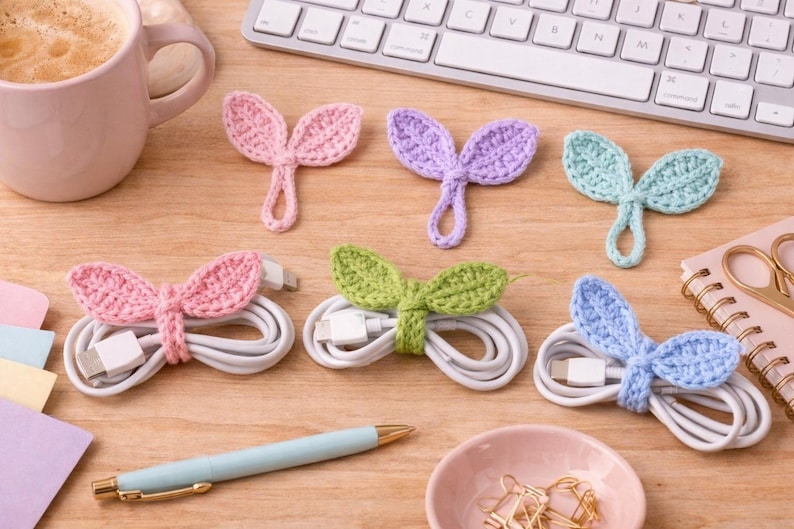 May include: A collection of colorful, handmade crochet cable organizers shaped like leaves. The organizers are in pink, purple, green, and blue, each wrapped around a white charging cable. A keyboard, coffee cup, and pen are also visible.